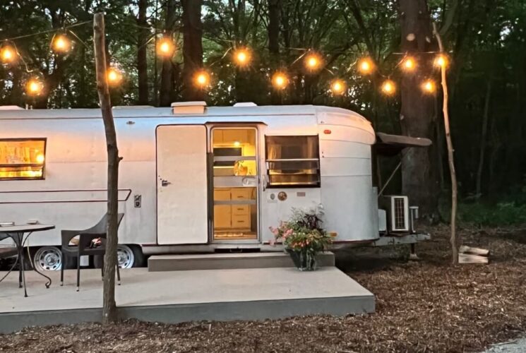 Large silver vintage airstream nestled in the woods, with patio holding table and two chairs and string lights in the trees