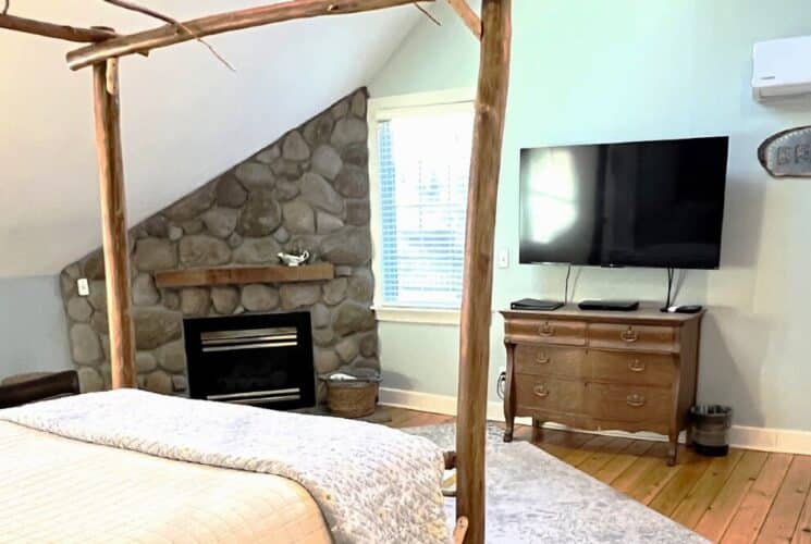 Bedroom with golden maple hand-built bed, stone fireplace, dresser with TV and wood floors.
