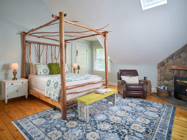 Bedroom with pine wood floors, a blue tapestry area rug, a golden maple hand-built bed, white nightstands with lamps on either side of the bed, leather chair and stone fireplace.