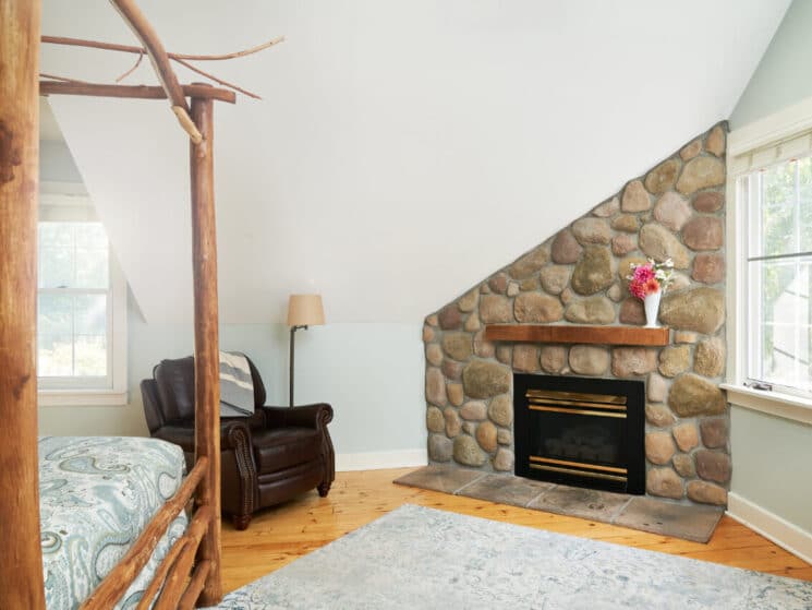 A bedroom with the corner of a bed with a wood canopy, pine wood floors, a light blue area rug, a brown leather chair next to a fireplace in a rock hearth.