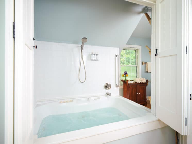 A jetted tub with bubbling water and a shower above it, with open doors along with a cupboard with rolled towels nearby.