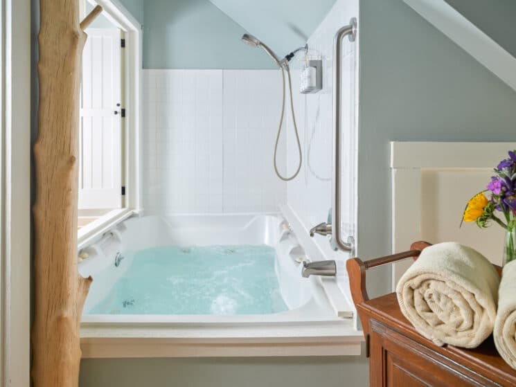 A jetted tub with bubbling water, an overhead shower, and a wood cupboard with tan towels on it along with a vase of colorful flowers.