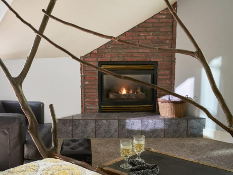Black leather chair next to a cozy gas fireplace with brick detail and 2 glasses of white wine on a trunk at the foot of a bed made of pieces of wood.