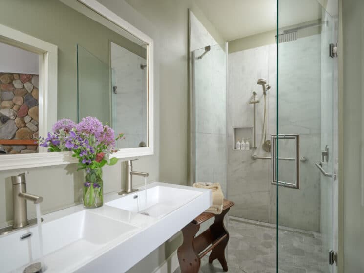 A modern bathroom with grey tiled floor, a shower with bth amenities, and a sleek double sink with a vase of purple flowers.