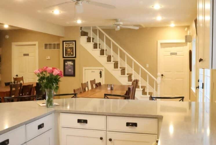 Large white kitchen with two large wood dining tables in front of a stone fireplace and a stairwell leading to a second floor