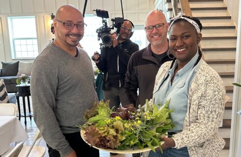 Two men with a woman holding a platter of leafy greens standing in front of a videographer