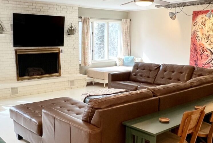 Large brown leather sectional in a spacious living room with large windows and TV over a white brick fireplace