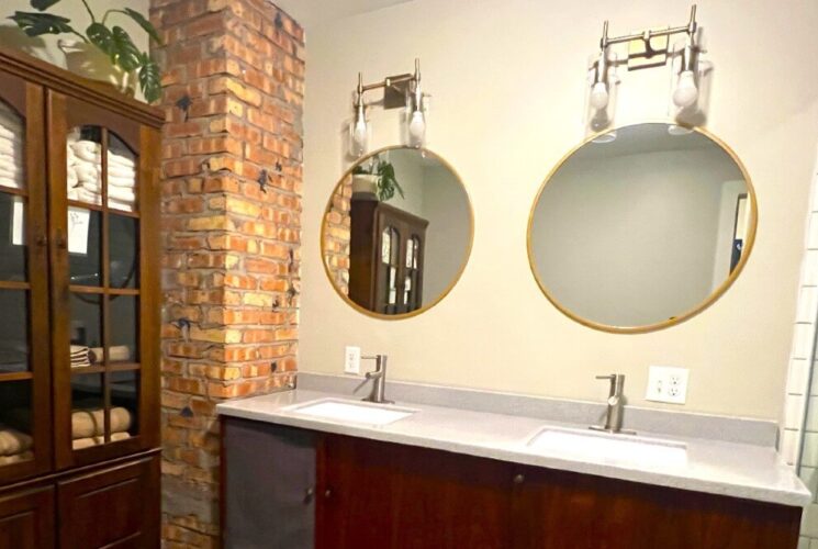 Elegant bathroom with two sink vanity, two round mirrors in gold trim, large hutch and tiled shower with glass door