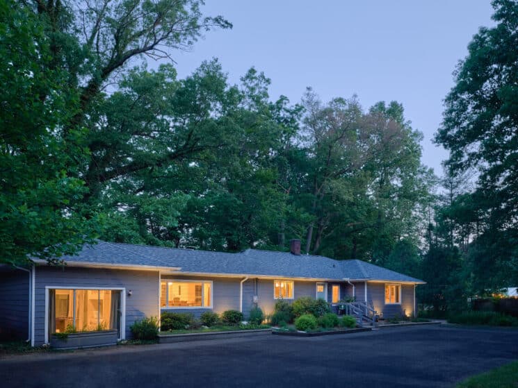 The exterior of a sprawling blue house lit up at dusk amidst mature trees.