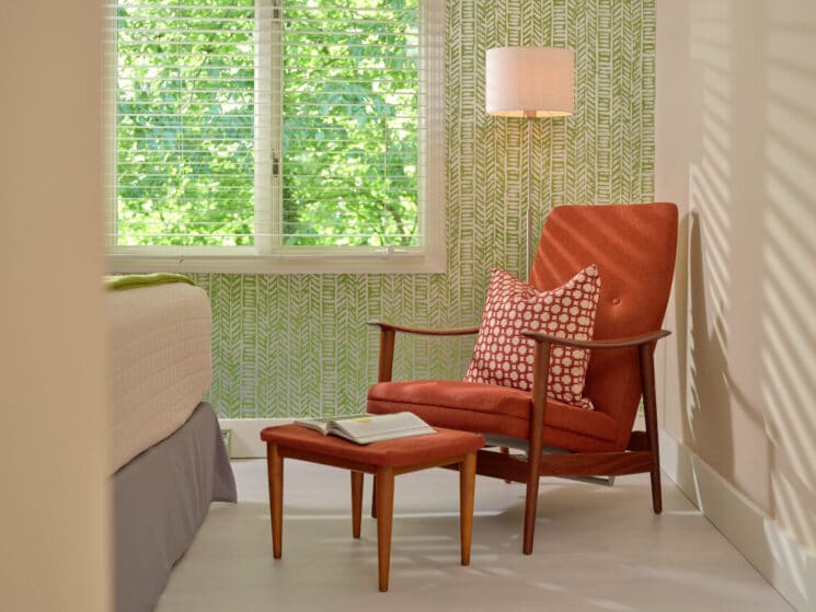 A corner of a bedroom with a orange chair and footstool with green wallpaper, the corner of a bed, and a window letting in bright light.