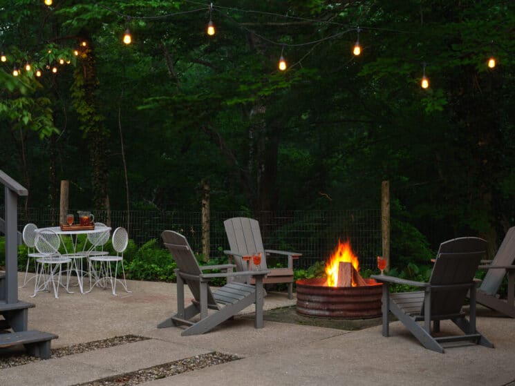 An outdoor patio at night with a roaring fire in a fire pit, 4 adirondack chairs around it, a white metal table and 4 chairs nearby, and twinkle lights hanging from trees.