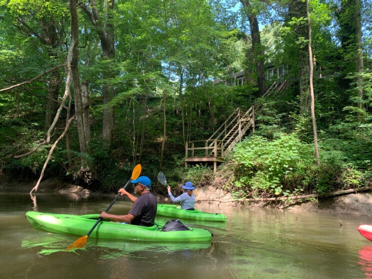 A couple kayaking in green kayaks on a river with a stairway heading up to a house above.