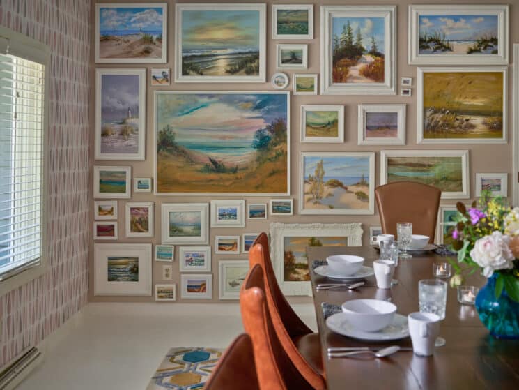 A dining room with a wood table set with white plates, bowls and coffee cups, a blue vase with fresh flowers, and a wall full of landscape pictures.