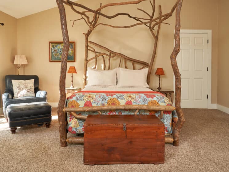 Elegant bedroom with a crooked four-poster bed made from sassafras branches, 2 leather chairs, a writing desk with lamp, cathedral ceilings, carpeted floors and slider door to a patio.