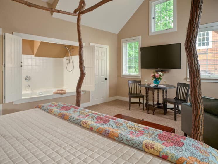 A spacious bedroom with the foot of a bed with sassafras branches as a canopy, a sitting area with a wood table and 2 chairs with a vase of flowers on it, and a flat screen TV above the table, and a bathtub built into the wall.