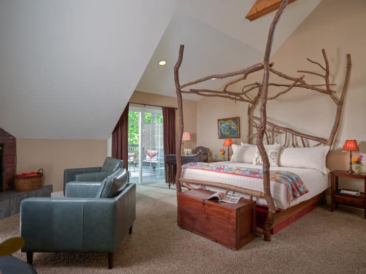 Elegant bedroom with four poster bed made from Sassafras wood branches, 2 club chairs, and a table with 2 glasses of white wine with sliding doors leading to an outside patio.
