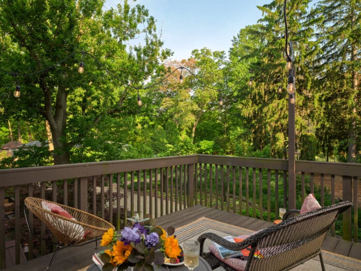 A wood deck with 2 wicker chairs, a small table in between them with a bouquet of colorful flowers and a glass of white wine, overlooking lush trees and grounds.