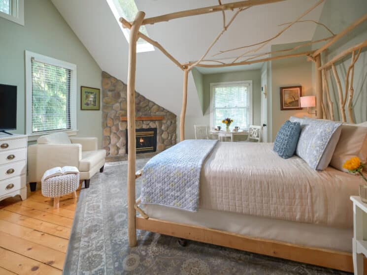 A bright and serene bedroom featuring a custom silver maple artisan-made bed, cathedral ceilings, stone fireplace, a table and chairs in front of a window, and wood floors