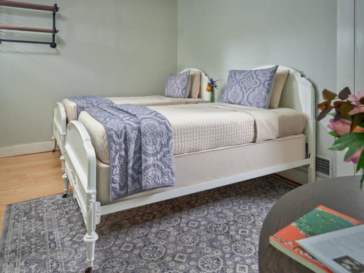 A bedroom with 2 twin beds with white headboards and footboards, cream colored bedding with blue accents, wood floors with a blue and white area rug, and a table with some books and a vase of flowers.