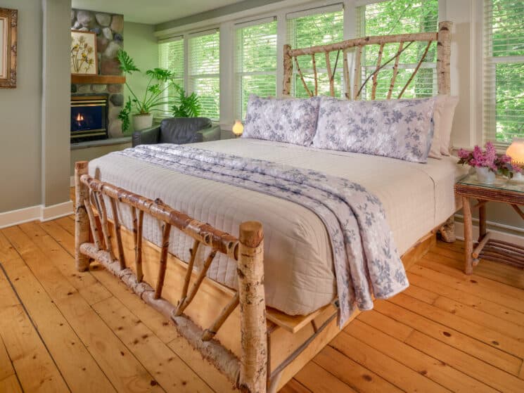 A bedroom with knotty pine floors, a birch bed with cream, blue and white bedding, birch nightstands with lamps on either side of the bed, a leather armchair net to a fireplace with a large plant in in a white pot.