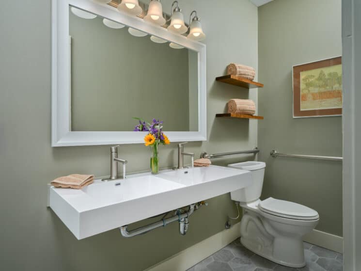 A bathroom with sage green walls, grey tiled floor, a chic vanity with 2 square sinks and a vase of purple and orange flowers, a toilet, and 2 wood shelves with rolled towels on them.