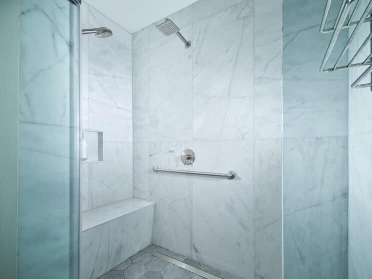 A white marble shower with dual shower heads and a sitting bench, along with a grab bar.