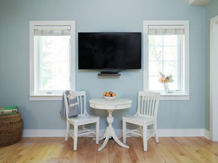 Small white table with two chairs beneath a TV on the wall flanked by two large windows