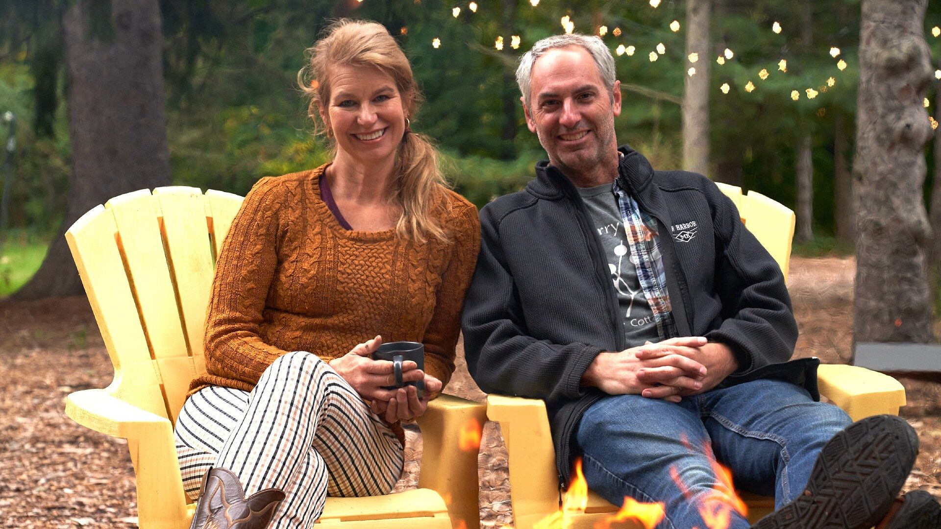 A man and woman sitting outdoors in yellow Adirondack chairs in front of a fire