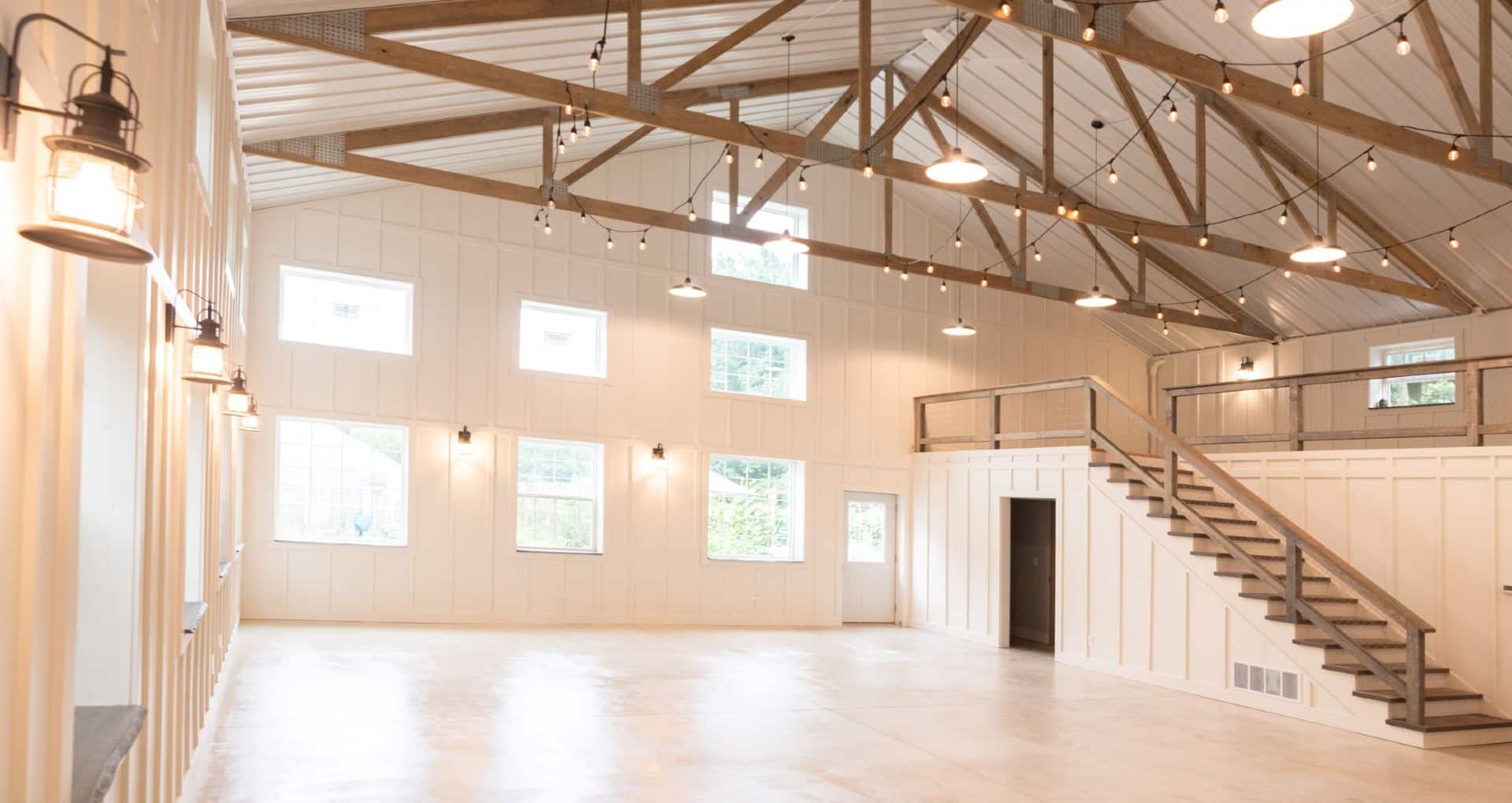 An empty large white barn with many bright windows, wood beams and string lights