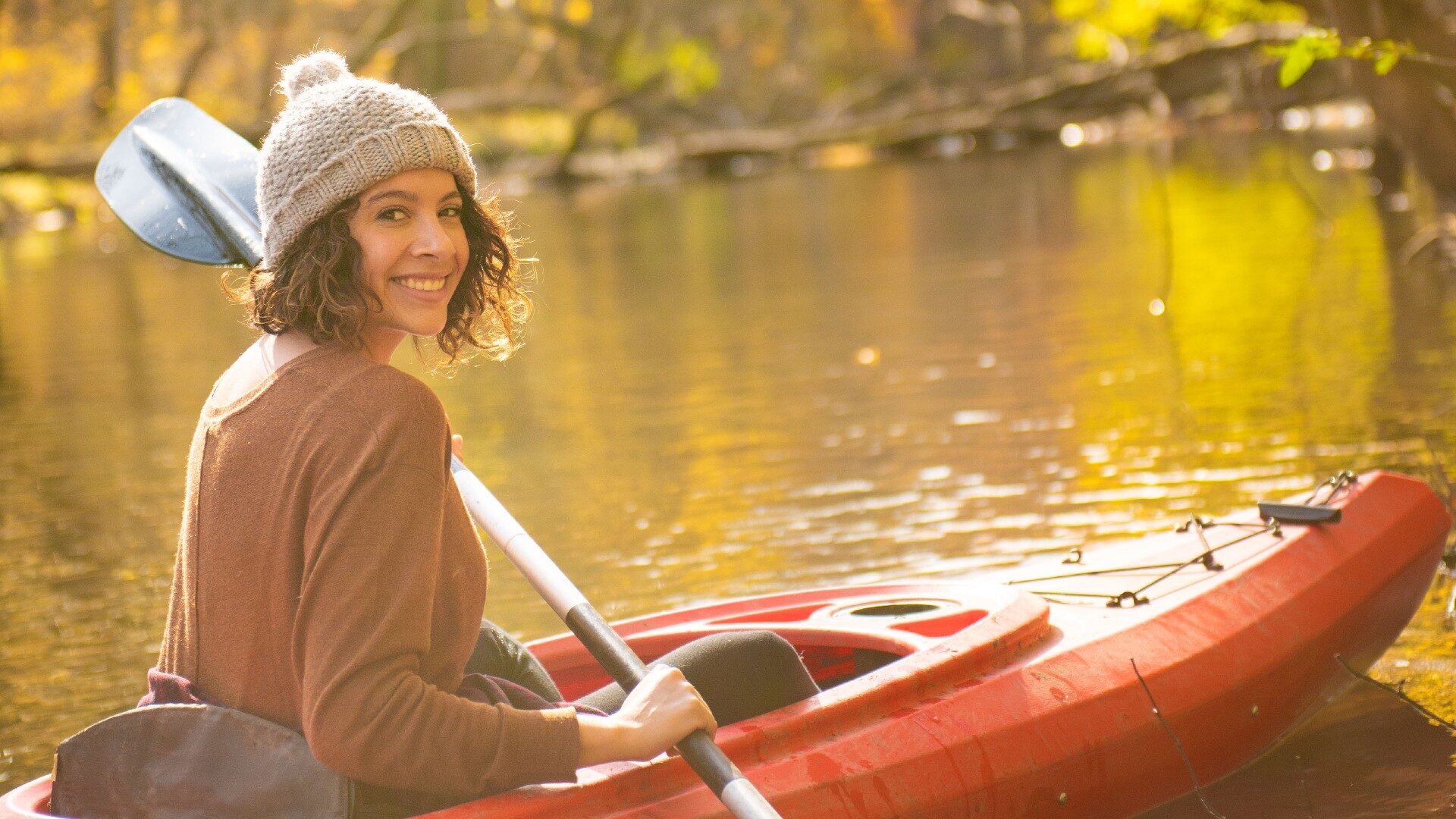 A girl in a snow hat kayaking in a red kayak down a creek bathed in sunlight
