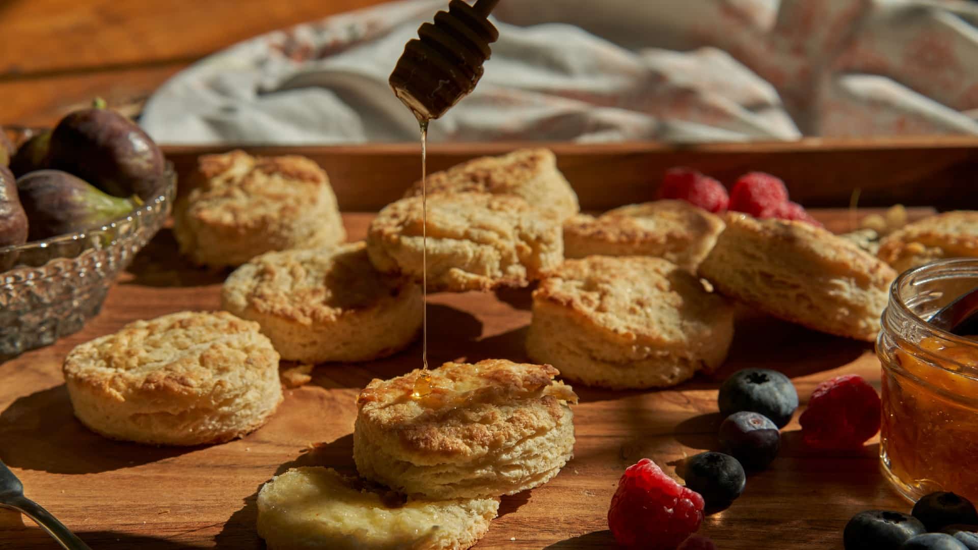 Freshly baked biscuits with honey being drizzled over top and berries on a wooden tray