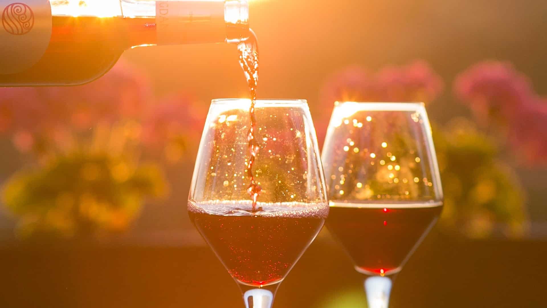 Red wine being poured into two wine glasses with flowers and golden sunshine in the background