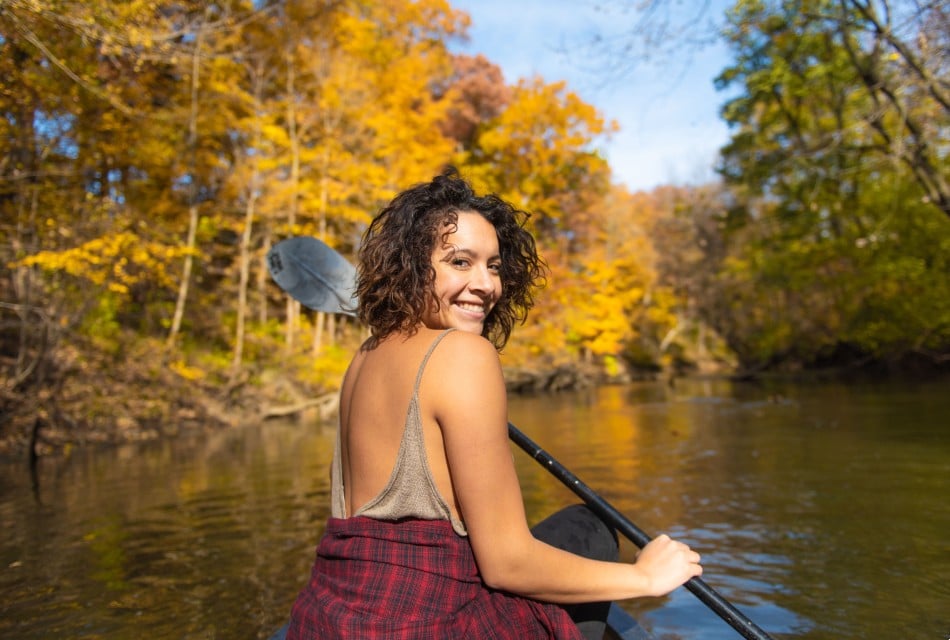 A woman with brown hair and smiling, kayaking in a pond surrounded by colored trees