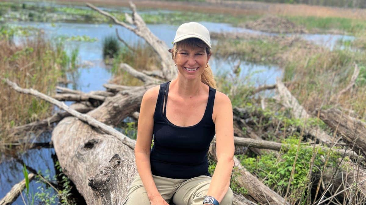 Smiling woman in a black tank top and beige cap sits on fallen logs by a marshy wetland.