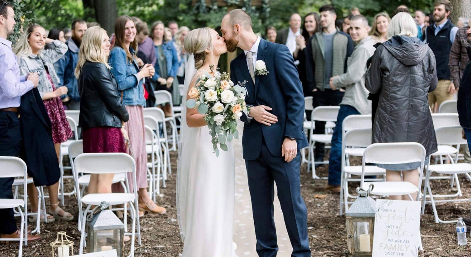 A bride and groom sharing a kiss at the end of the aisle with their guests smiling behind them