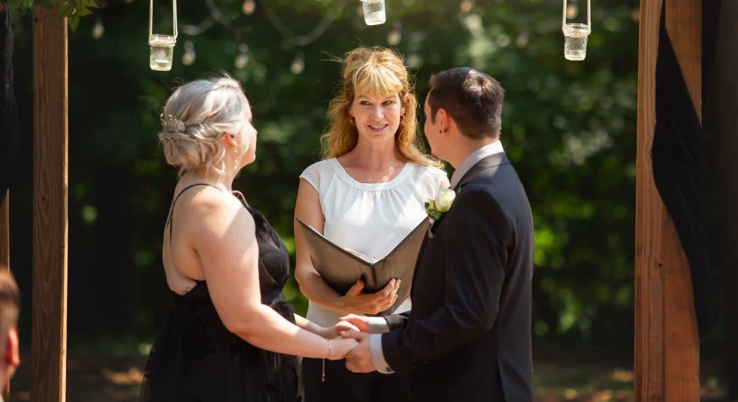 A bride and groom standing outdoors under a wooden arch in front of a female officiant