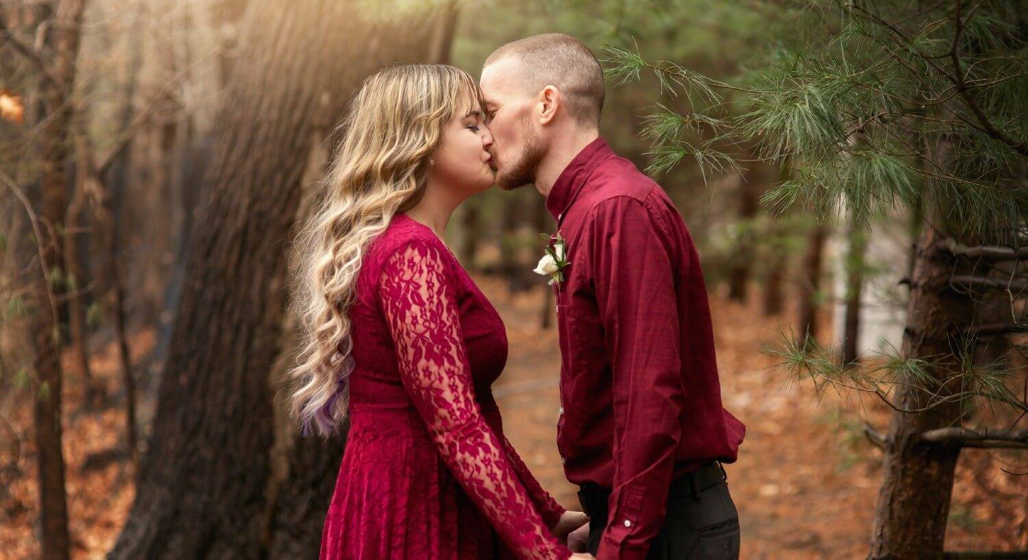 A man and woman both wearing burgundy sharing a kiss in the woods while holding hands