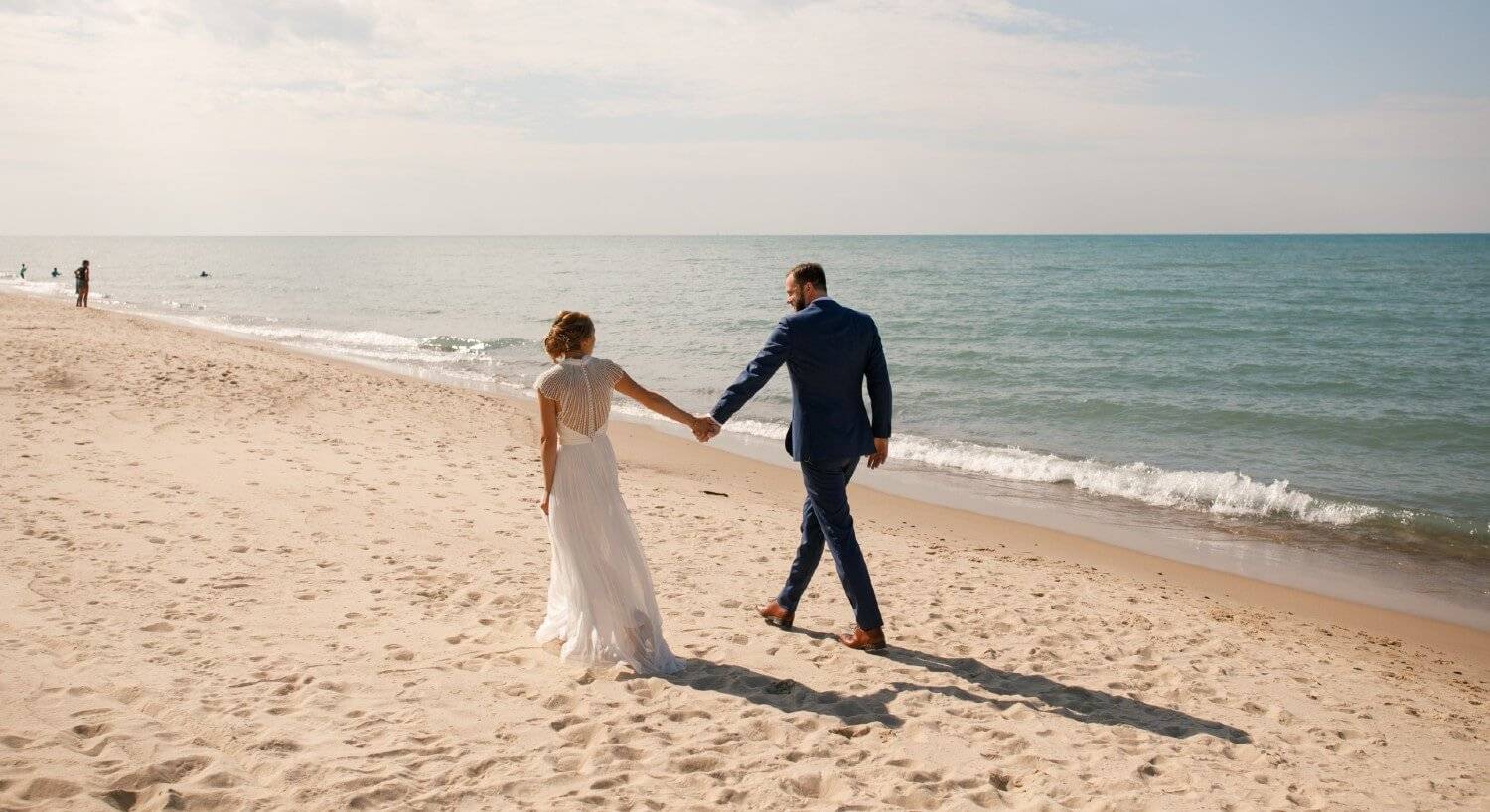 A bride and groom walking hand in hand down a beach next to the water