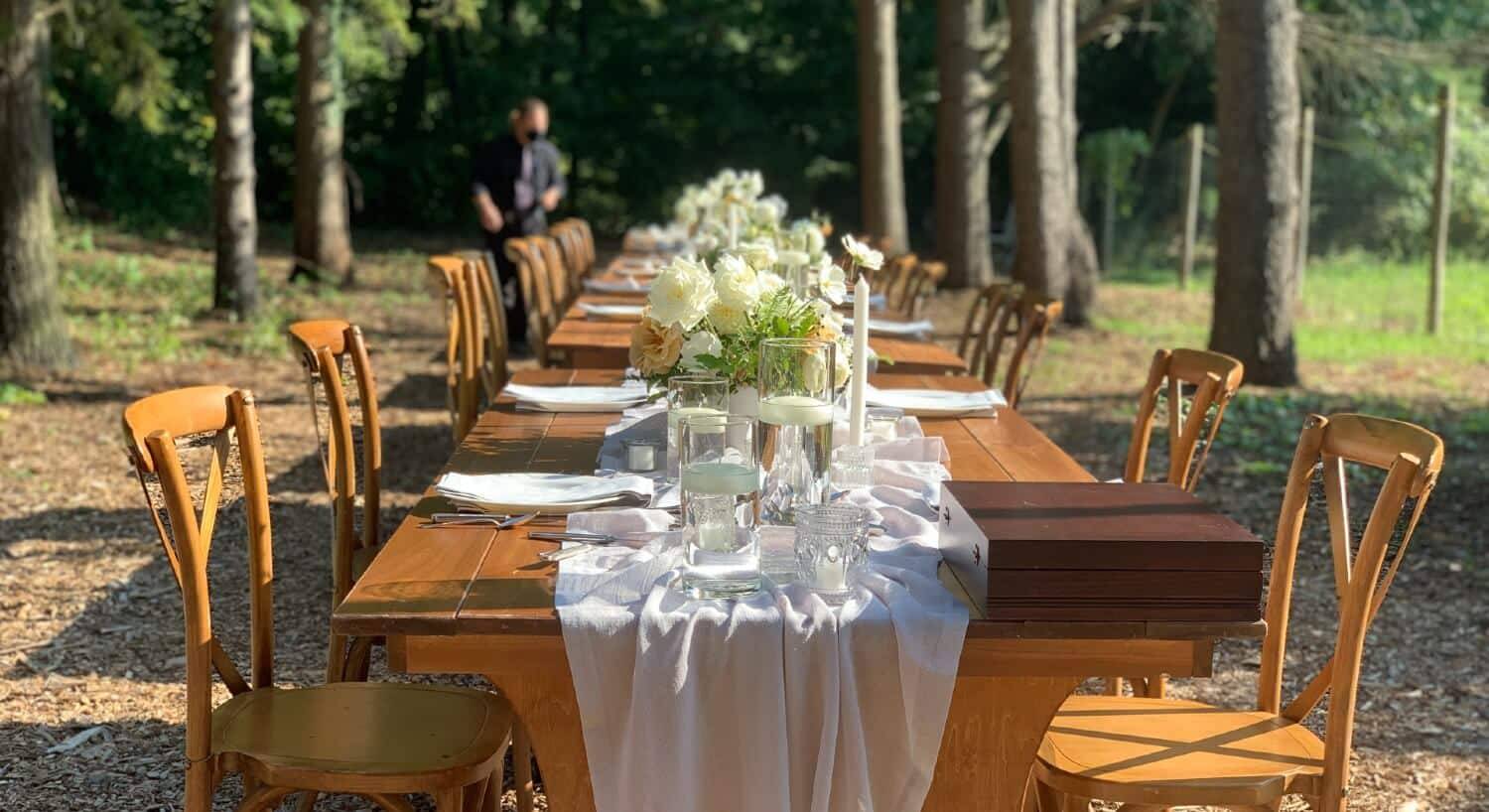 A long wood table set up outside with a white runner and several vases of white flowers