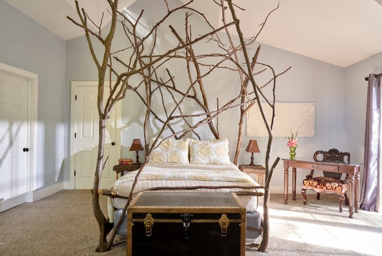 Bright and cozy bedroom with custom queen bed with rustic branch details, chest and writing desk near a large window with curtains