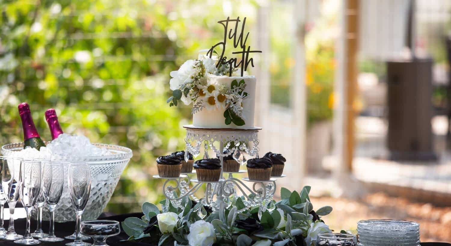 A wedding cake and chocolate cupcakes on a table with two champagne bottles on ice with champagne flutes