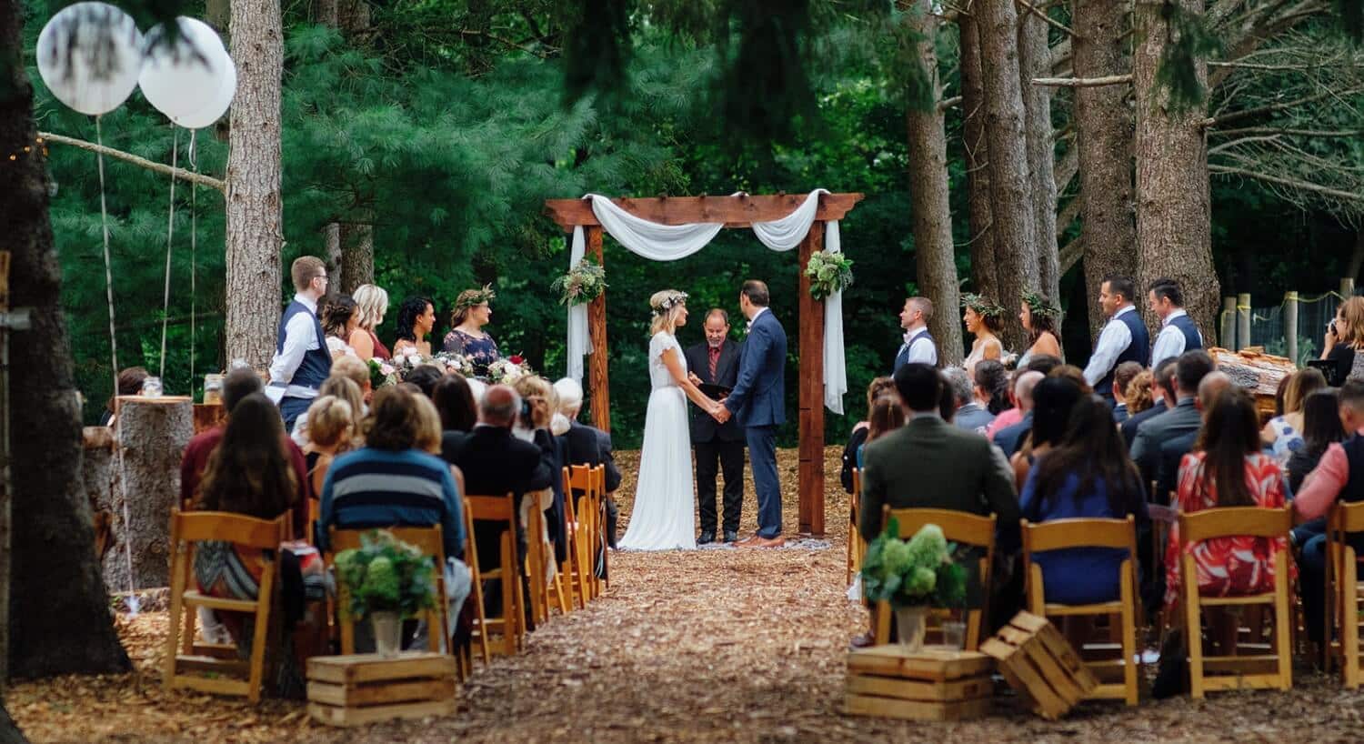A bride and groom standing under a wooden arch with a small group of guests surrounded by woods